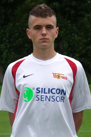 Ein junger Fußballspieler in einem weißen Trikot mit roten Akzenten und Sponsorenlogo steht auf einem Platz.