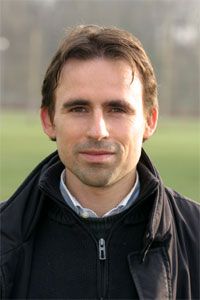 Porträt eines Mannes mit dunklem Haar, der einen schwarzen Mantel trägt, vor einem verschwommenen Hintergrund im Freien.