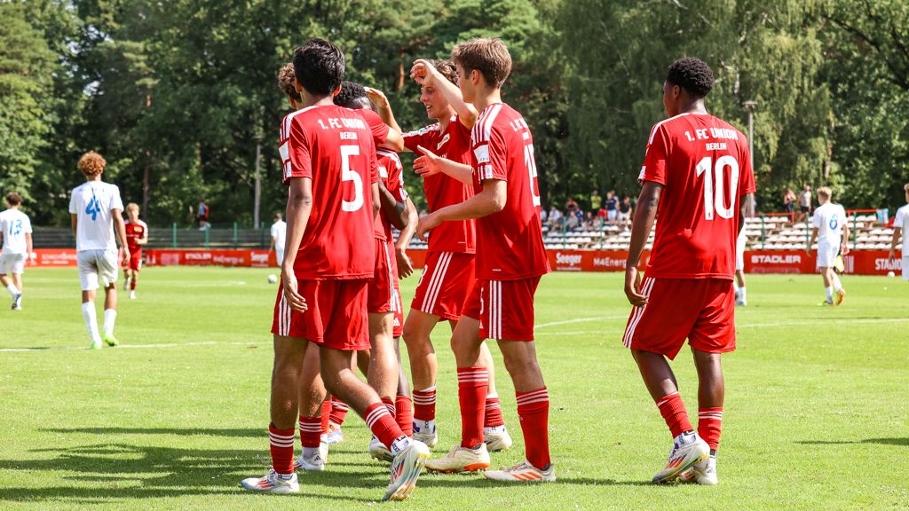Spieler in roten Trikots feiern einen Torerfolg auf dem Fußballplatz. Im Hintergrund ist ein leeres Stadion zu sehen.