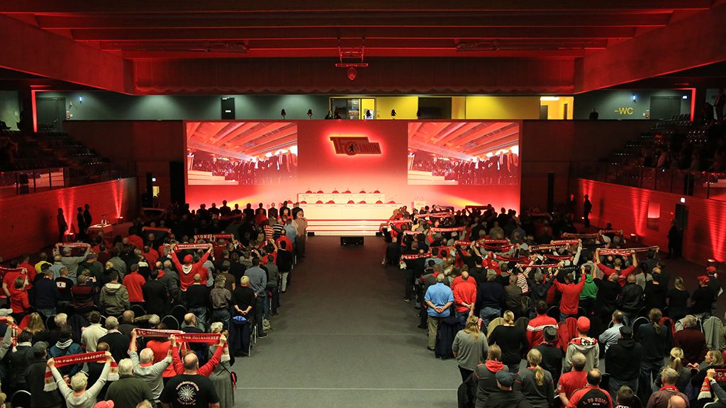 Menschenmenge in einem Veranstaltungsraum mit roten Lichtern, die Schals halten und auf eine Bühne mit einer Präsentation blicken.