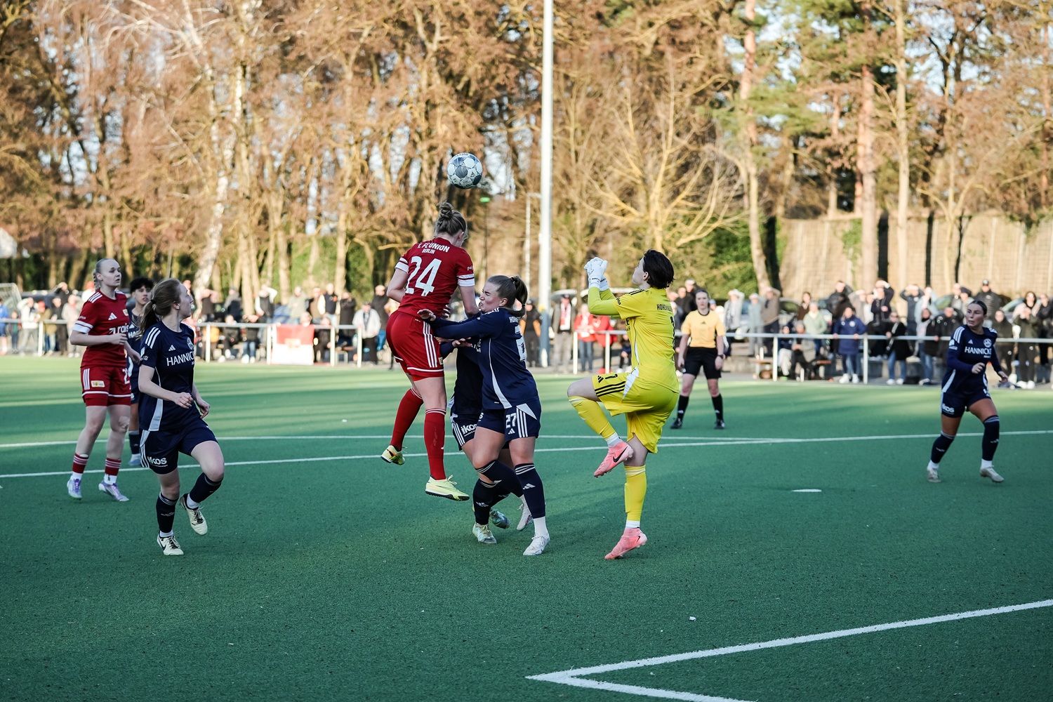 Eine Fußballspielszene mit einer Spielerin in rotem Trikot, die im Sprung einen Ball spielt, während die Torhüterin versucht, ihn zu fangen.