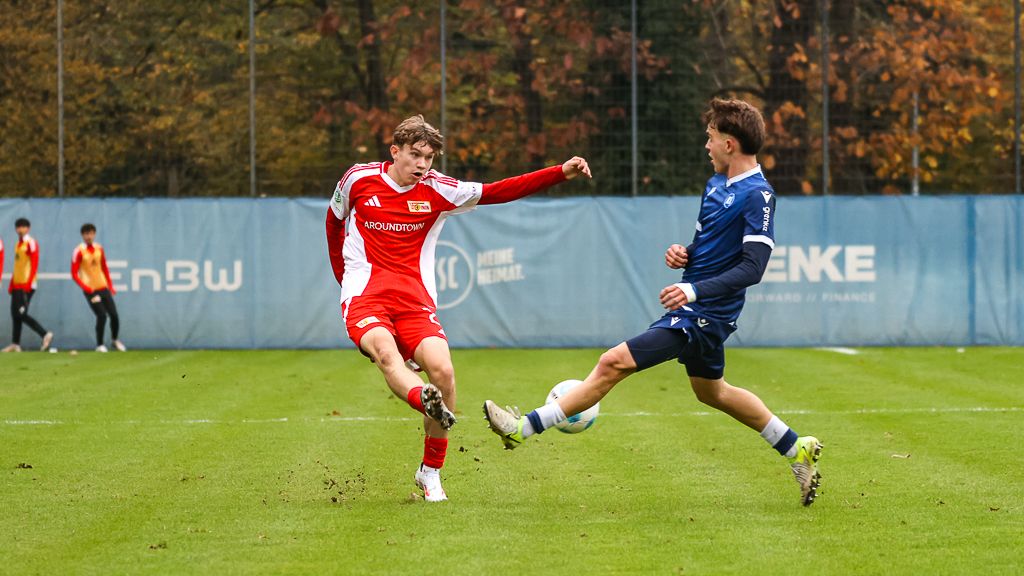 Zwei Fußballspieler im Wettkampf um den Ball auf einem grünen Platz. Im Hintergrund sind Zuschauer und eine Werbebande zu sehen.