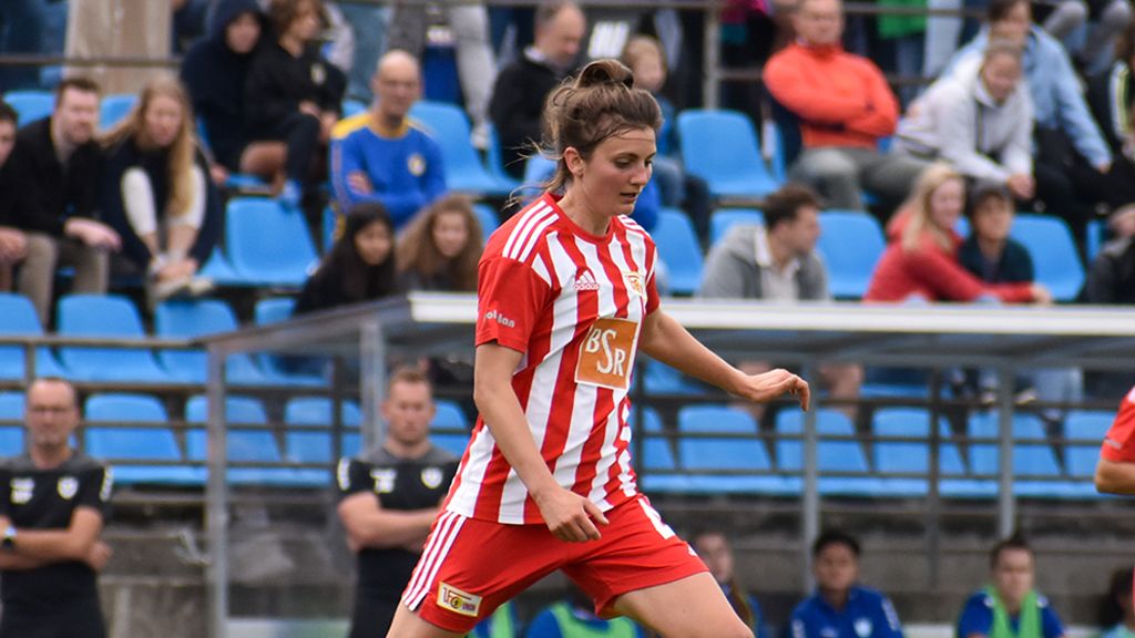 A female soccer player in a red-and-white jersey sprints across the field, with spectators in the background.