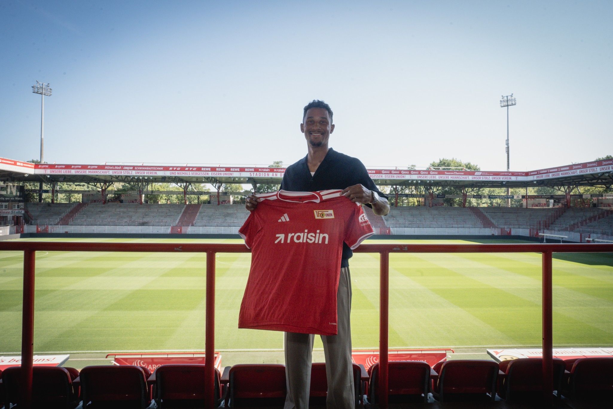 Ein Spieler hält ein rotes Trikot mit der Aufschrift "raisin" vor einem leeren Stadion mit Rasenplatz.