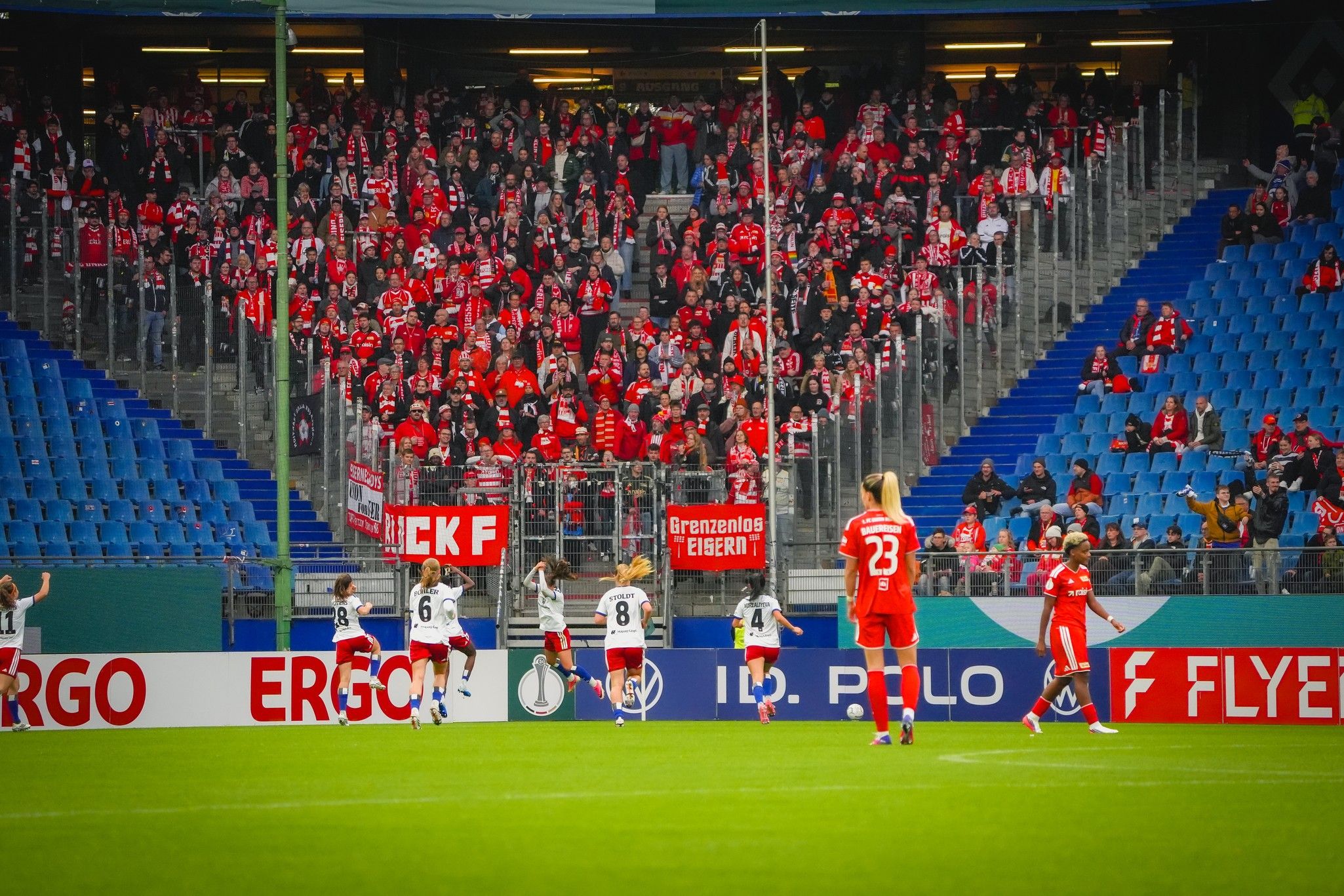Fans in rot-weißen Trikots jubeln im Stadion, während Spieler in weißen und roten Trikots auf dem Spielfeld agieren.