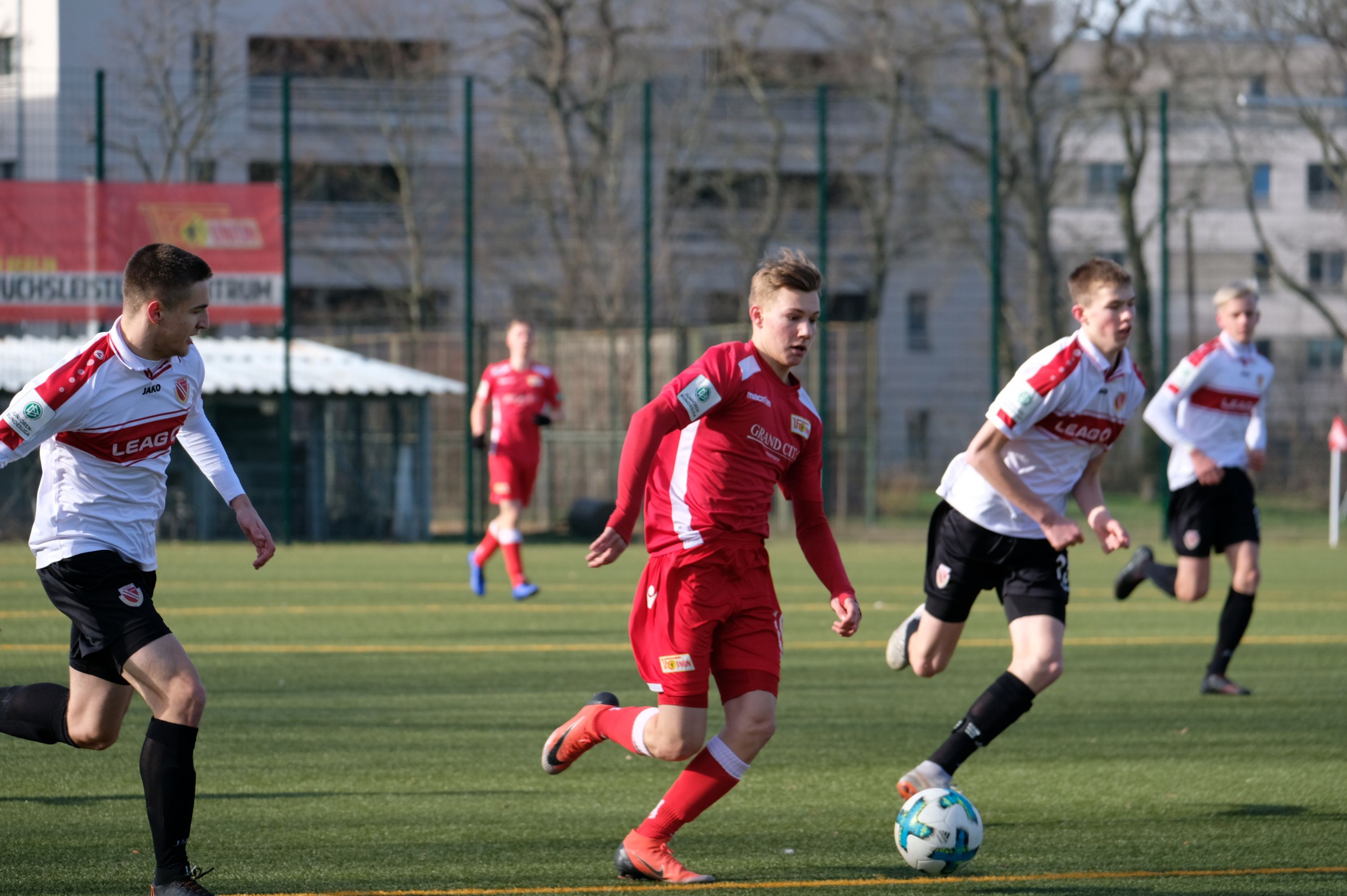Zwei Fußballspieler in roten und weißen Trikots laufen auf einem Kunstrasenplatz, während sie einem Ball nachjagen.
