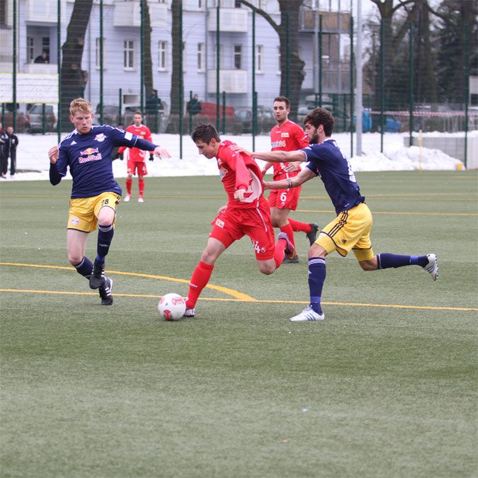Ein Fußballspieler in rotem Trikot dribbelt mit dem Ball, während zwei gegnerische Spieler in gelben und blauen Trikots versuchen, ihn zu stoppen.