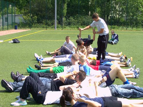 Eine Gruppe von Jugendlichen liegt auf einem Sportplatz, während ein Trainer Anweisungen gibt.