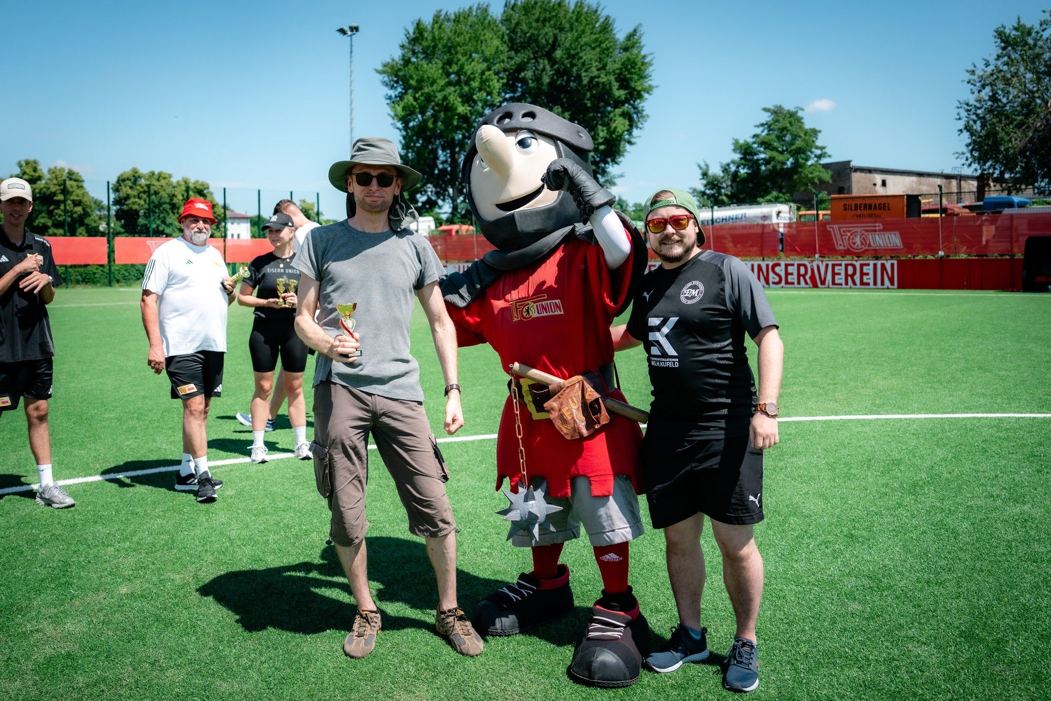 Zwei Männer posieren neben einem Maskottchen auf einem Fußballplatz, während andere im Hintergrund stehen.