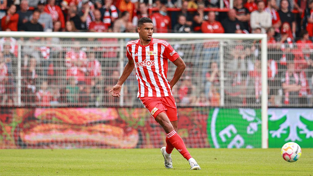 Fußballspieler in rot-weißem Trikot setzt zu einem Schuss an, im Hintergrund Fans in Stadion.