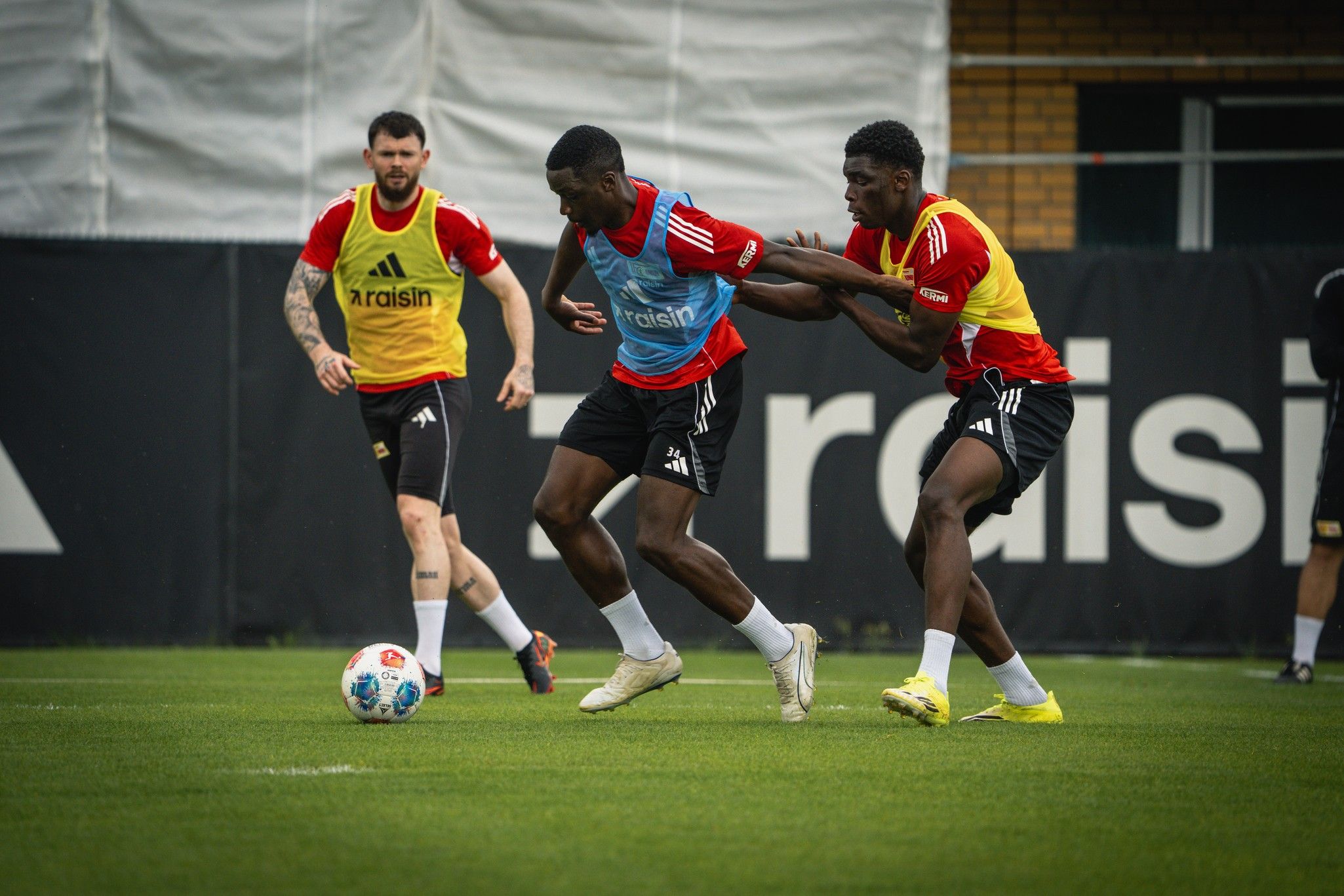 Drei Fußballspieler trainieren auf einem grünen Platz, zwei Spieler im Wettlauf und einer beobachtet.