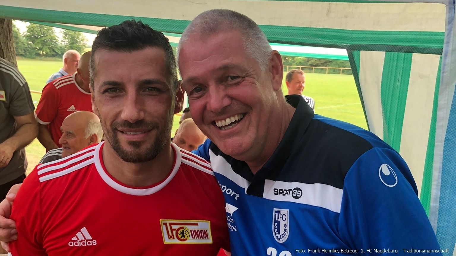 Zwei Männer lächeln in Sportbekleidung unter einem Vordach, im Hintergrund sind weitere Personen und ein Fußballfeld sichtbar.