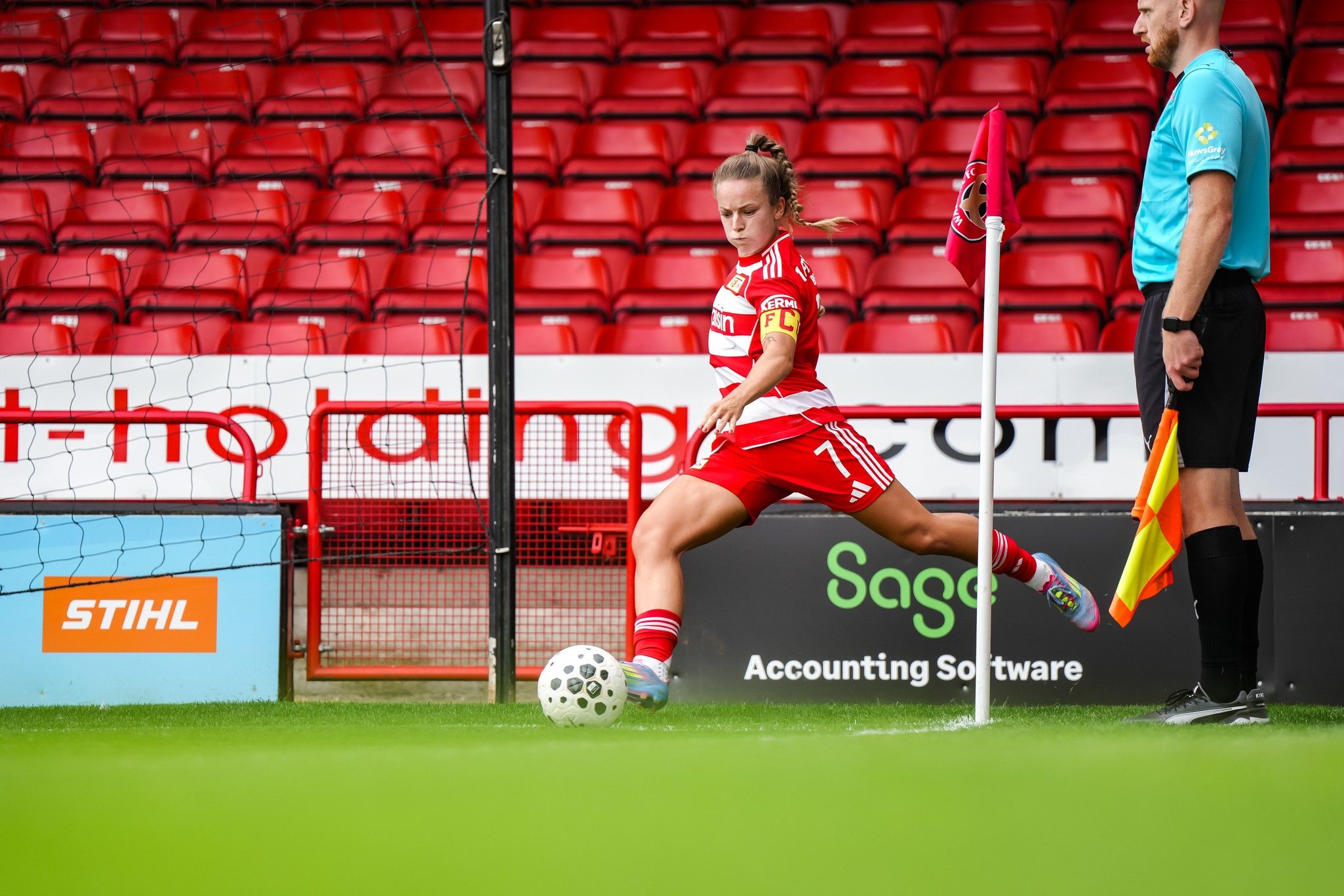 Eine Fußballspielerin mit Kapitänsbinde steht an der Eckfahne und führt einen Eckball aus. Ein Schiedsrichter steht neben ihr.