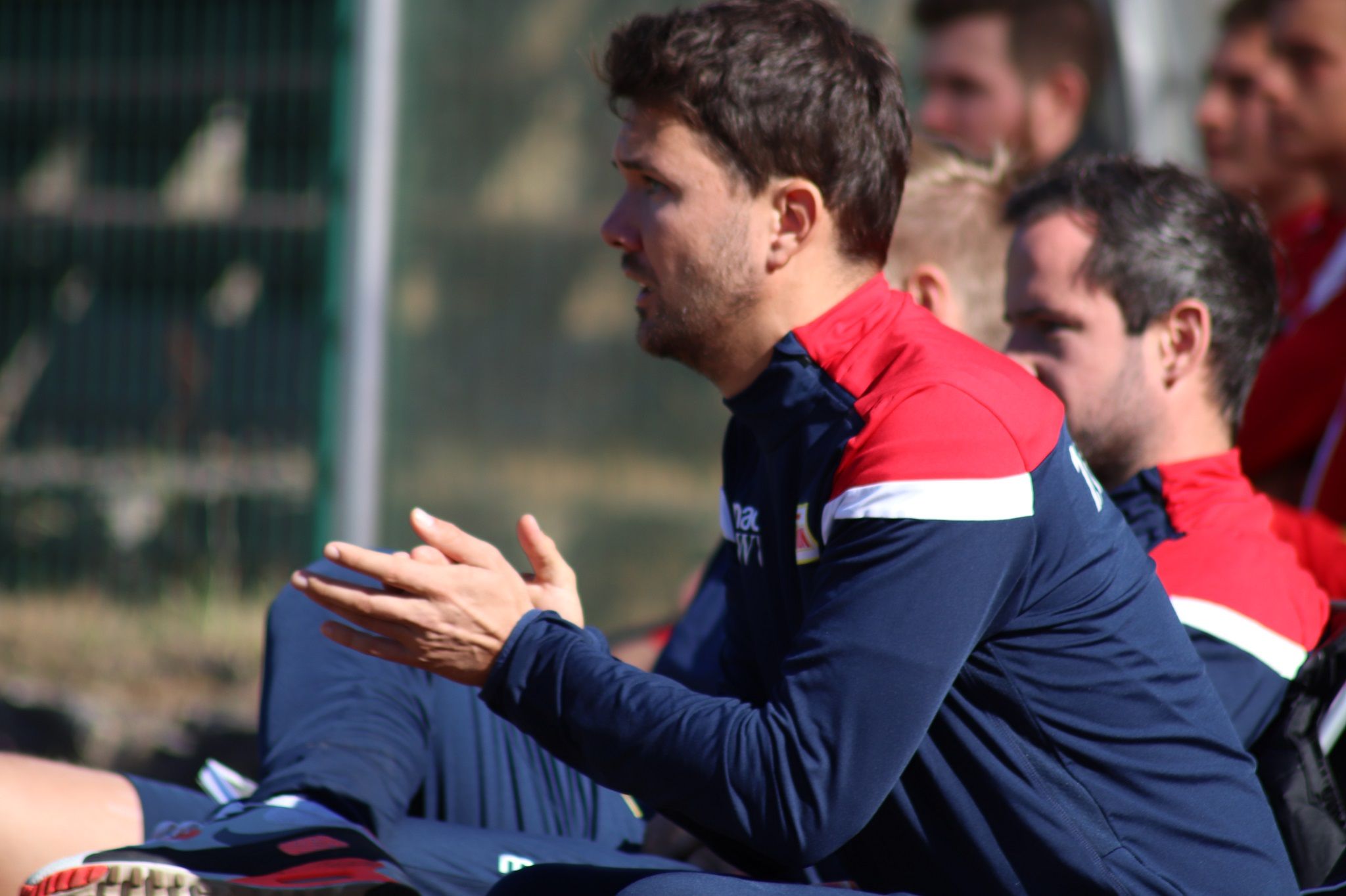 Ein Trainer sitzt an der Seitenlinie und clapt begeistert, während Spieler im Hintergrund auf der Bank sitzen.