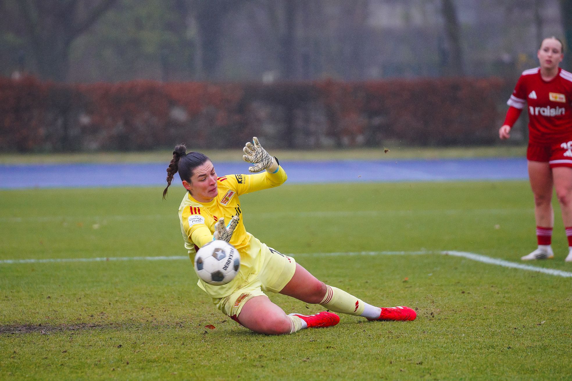 Eine Torhüterin in gelber Ausrüstung fängt einen Ball auf einem nassen Fußballfeld, während eine Spielerin im Hintergrund zusieht.