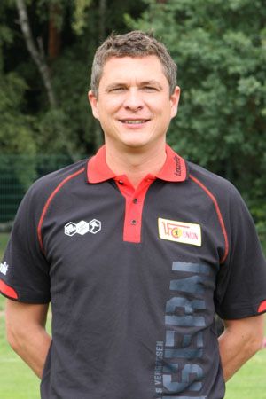 Ein lächelnder Mann in einem schwarzen Poloshirt mit einem roten Kragen, steht in einer grünen Umgebung.