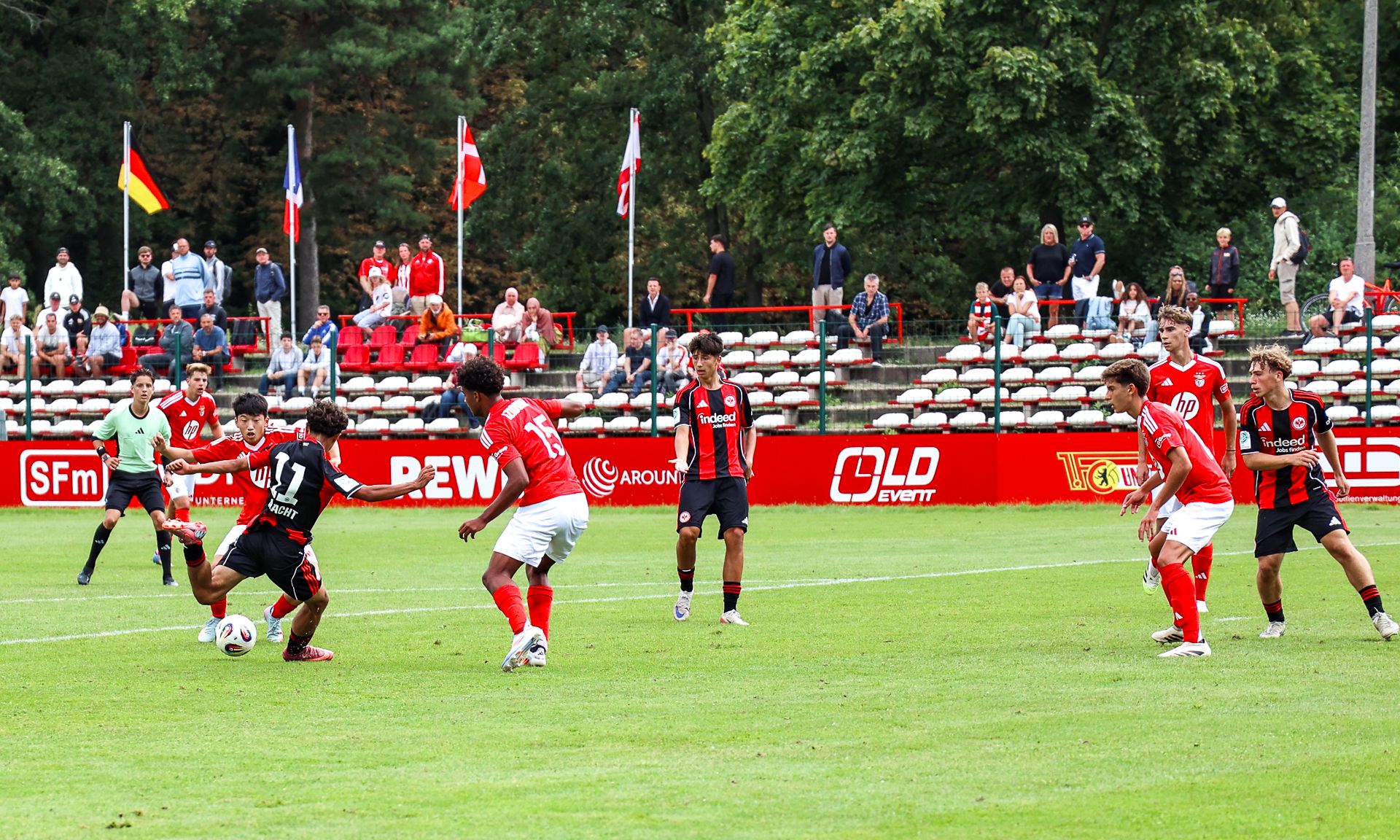 Spielszene auf einem Fußballfeld mit Spielern in roten und schwarzen Trikots, Zuschauer im Hintergrund, eine angespannte Spielsituation.
