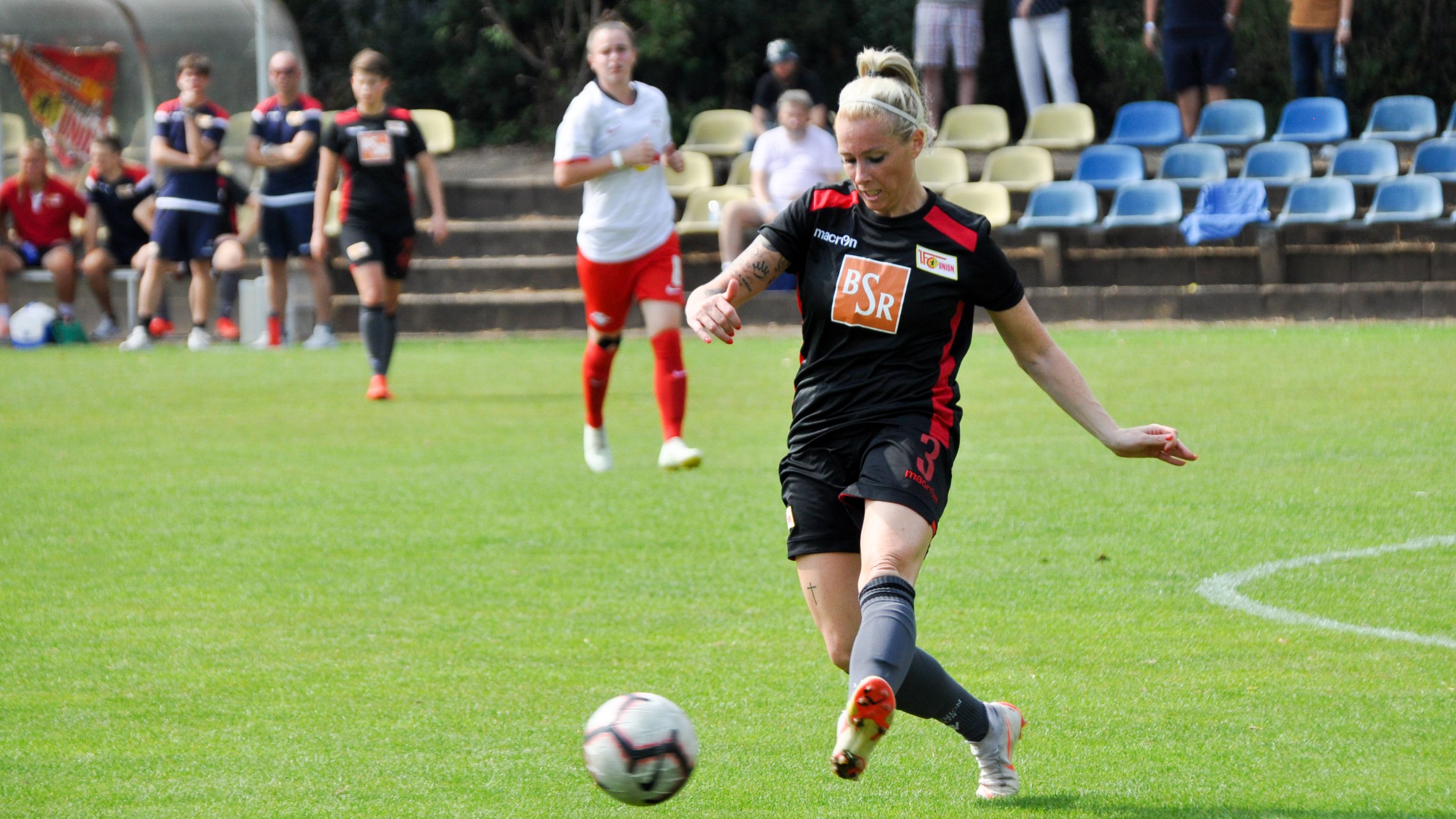 Eine Fußballspielerin in schwarzem Trikot tritt den Ball auf einem Rasenplatz, umgeben von anderen Spielern und Zuschauern.