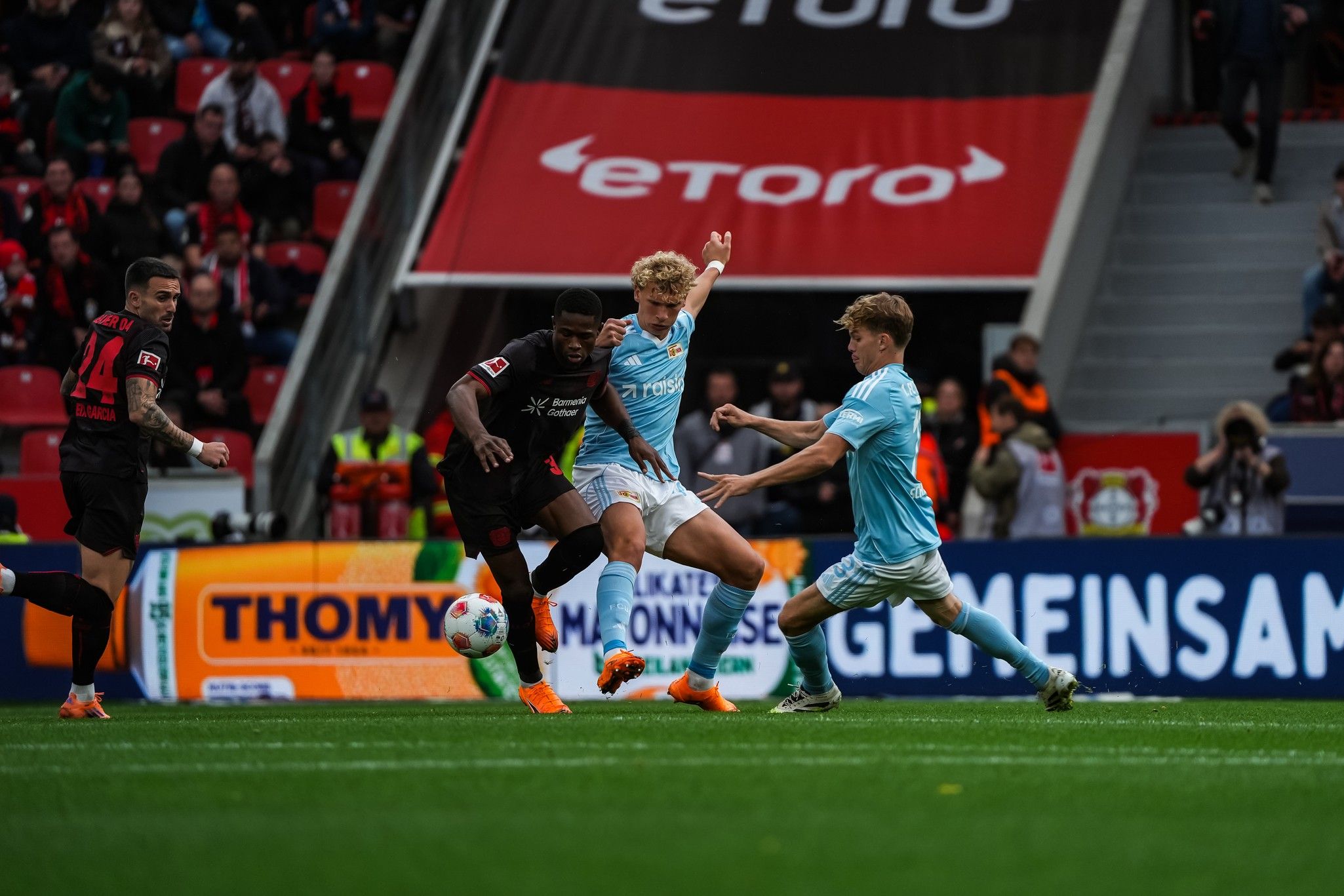 Fußballspieler im Duell auf dem Platz, hinter ihnen Zuschauer in einem Stadion, mit Werbebannern im Hintergrund.