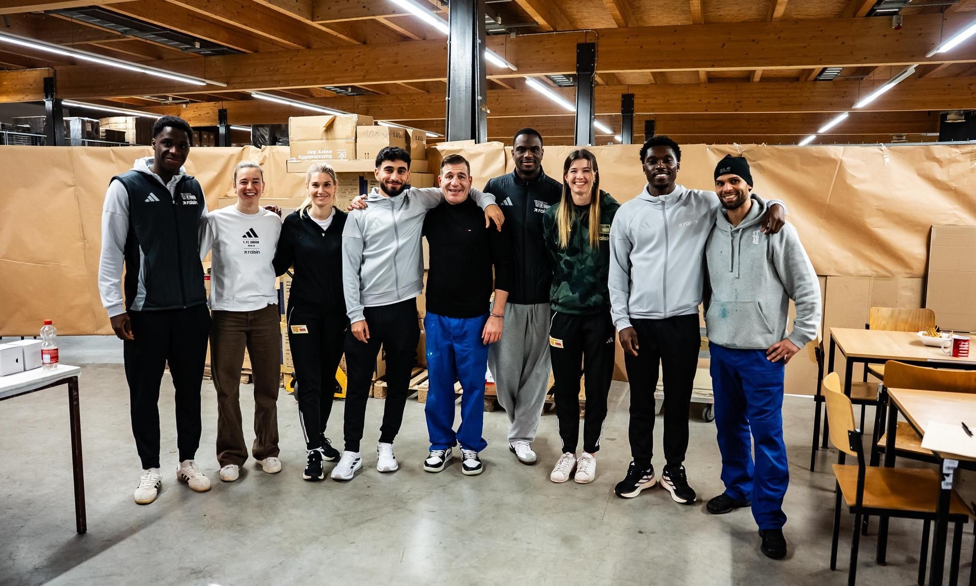 Eine Gruppe von neun Personen, die in Sportkleidung gekleidet sind, posiert gemeinsam in einem Lagerhaus.