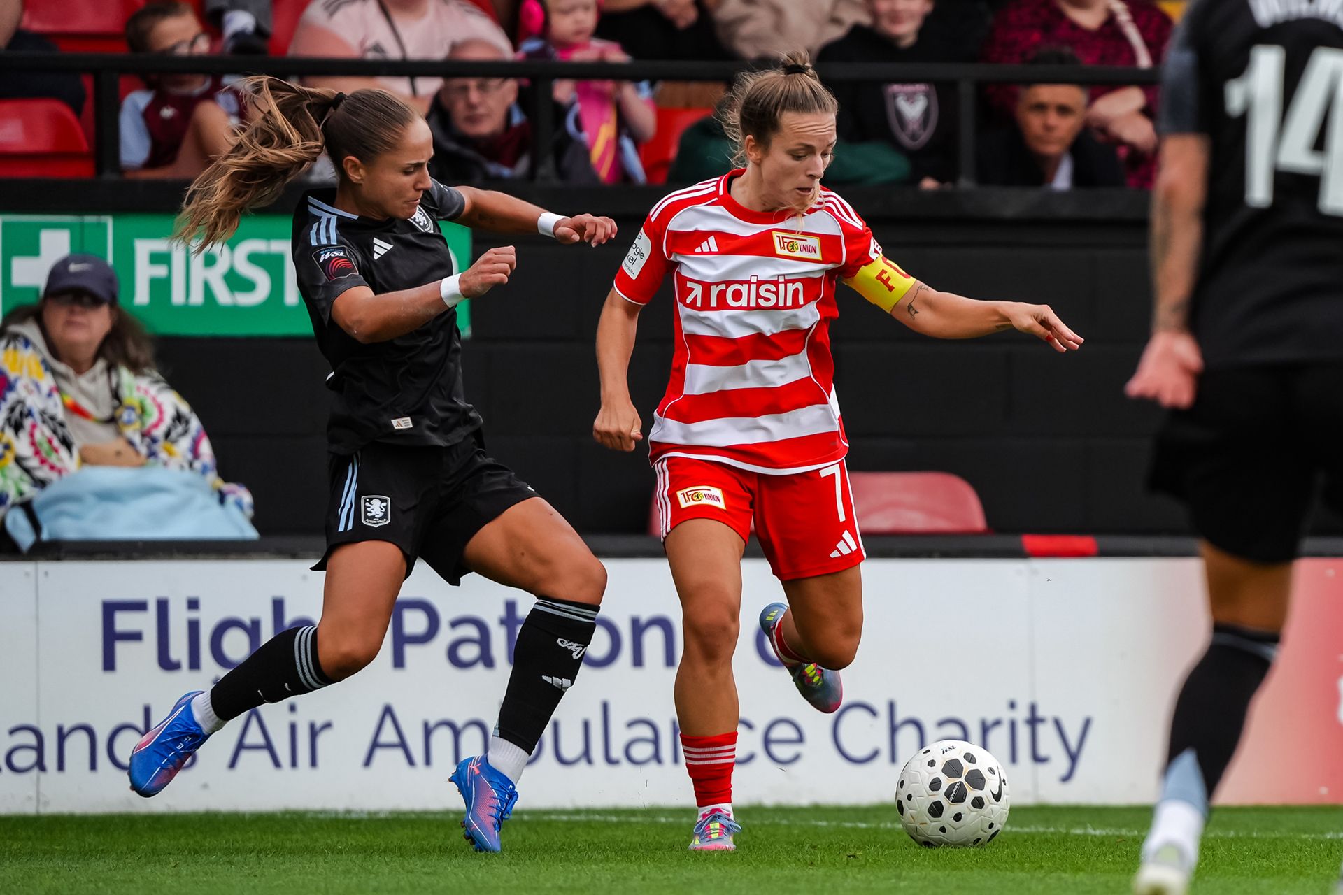 Zwei Frauenfußballspielerinnen im Wettkampf um den Ball in einem Stadion. Die eine trägt rot-weiße Streifen, die andere schwarz.