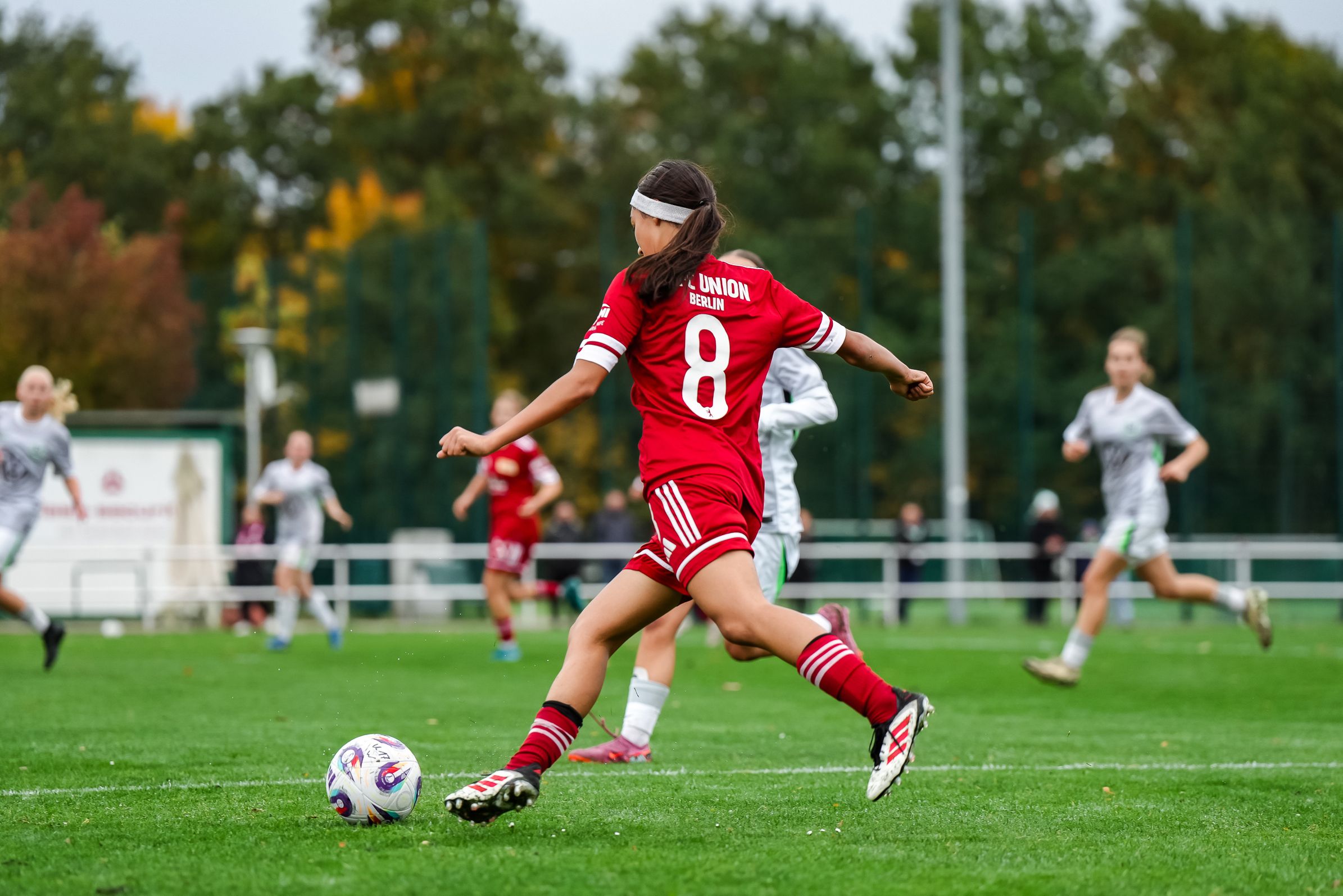 Eine Spielerin in rotem Trikot tritt den Ball auf einem Fußballfeld, während andere Spielerinnen im Hintergrund aktiv sind.