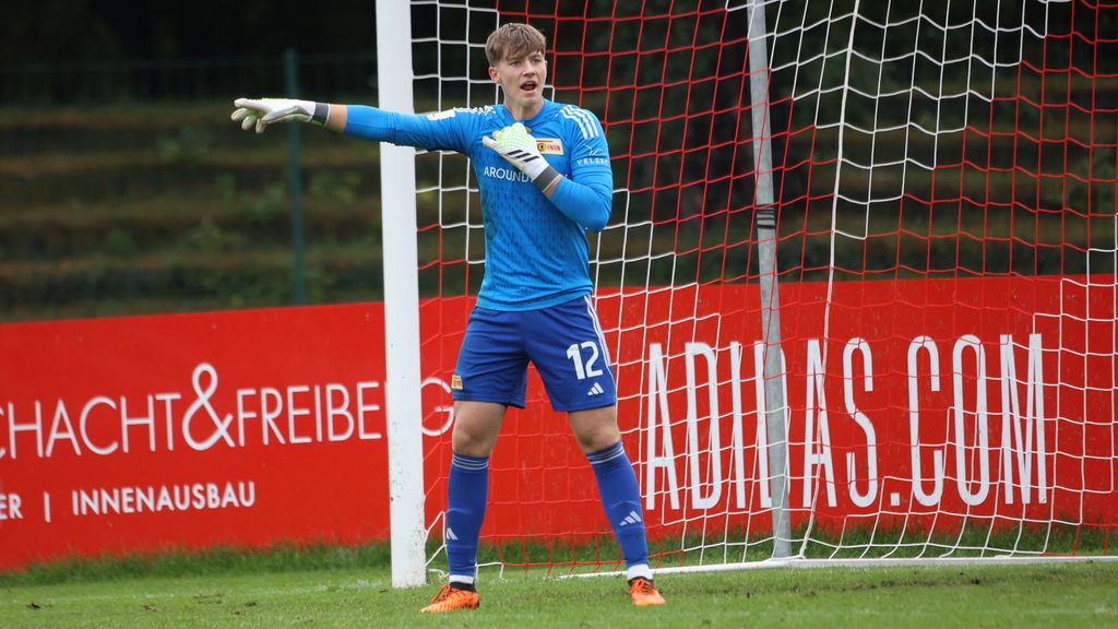 Ein Torwart in blauer Spielkleidung steht im Tor und gibt Anweisungen, während ein Fußballspiel stattfindet.
