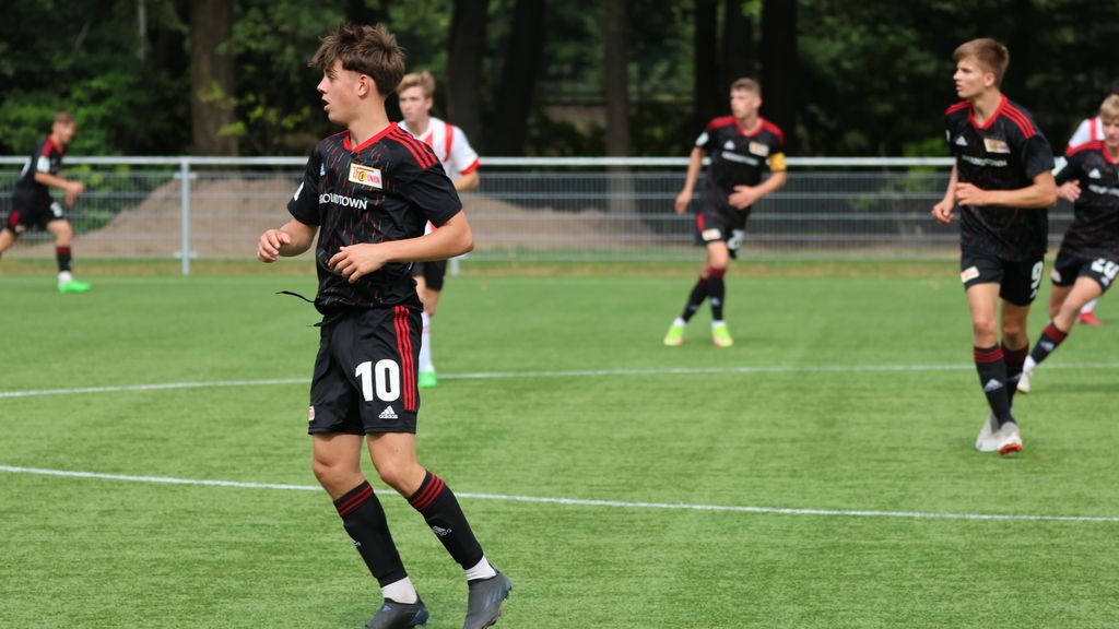 Ein junger Fußballspieler in schwarzem Trikot mit der Nummer 10 auf einem Rasenplatz, im Hintergrund weitere Spieler in Aktion.