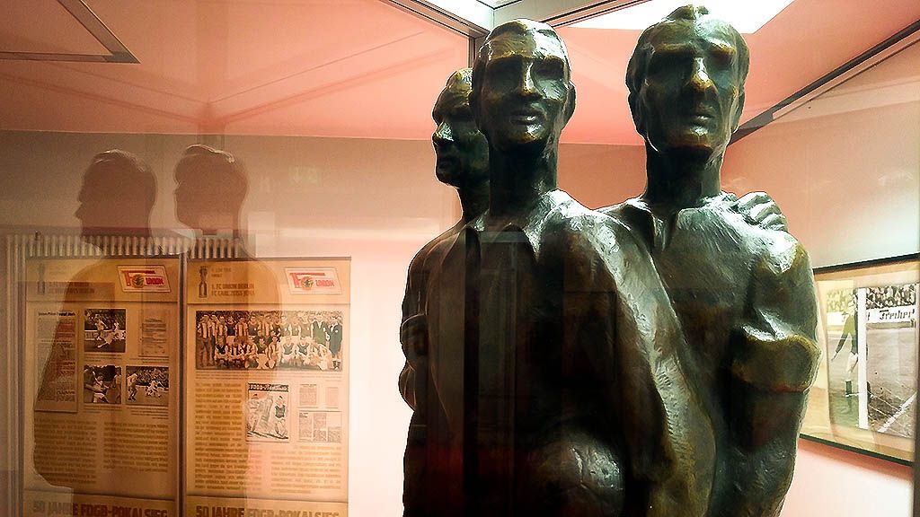 Bronzeskulptur von drei Männern, die sich aneinander lehnen, im Hintergrund Dokumente und Bilder an einer Wand.