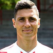 Ein lächelnder Fußballspieler in einem rot-weißen Trikot vor einer unscharfen Stadionkulisse.