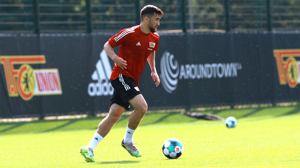 A soccer player in a red jersey is running on a training field with green grass and advertising banners in the background.