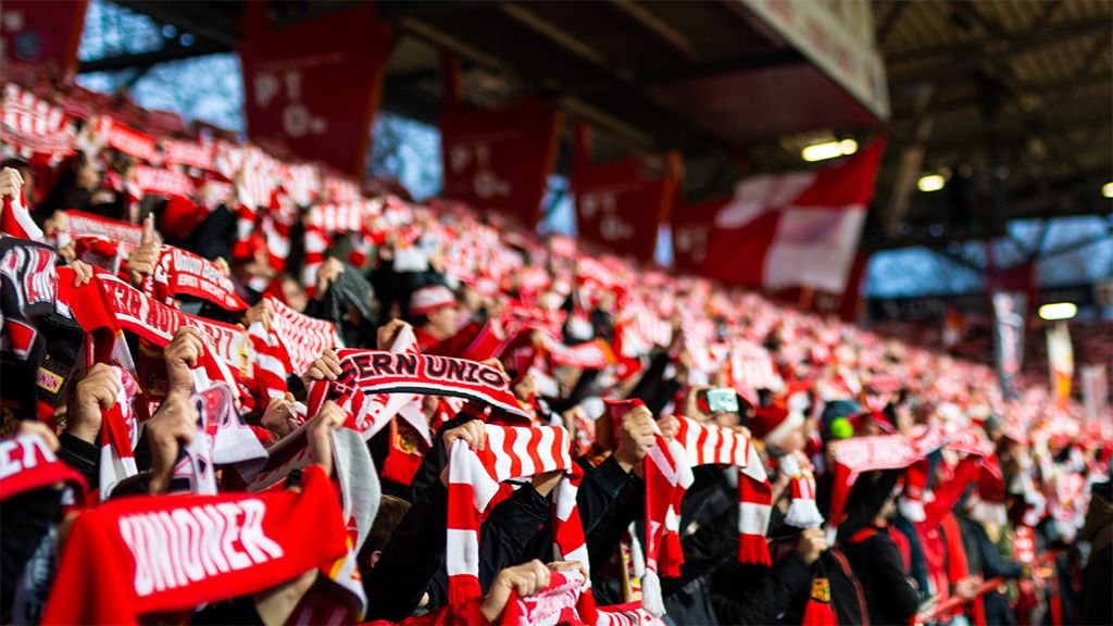 A large crowd in a stadium holding red and white scarves high and cheering for their team.