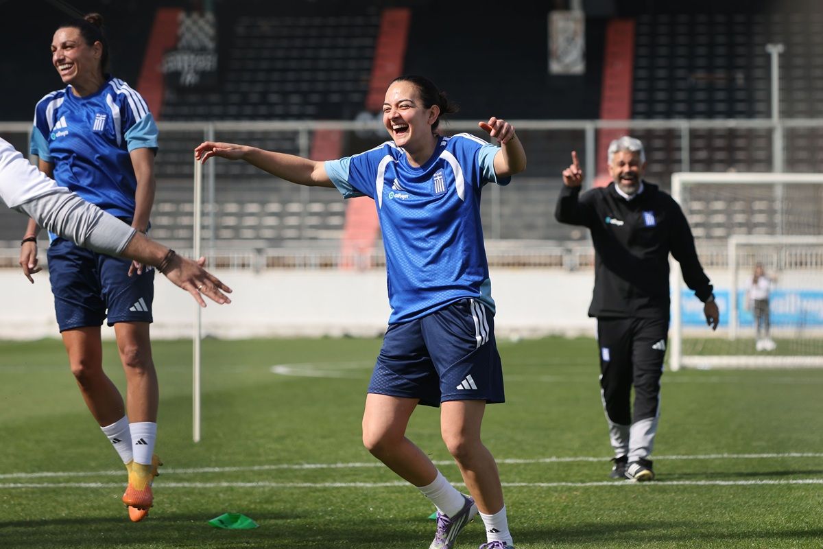 Eine Fußballspielerin in blauer Trainingskleidung lacht und feiert auf dem Spielfeld, während andere im Hintergrund zuschauen.