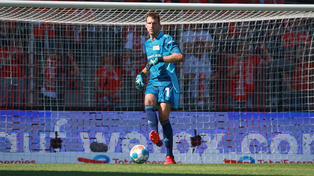 Ein Torwart in blauer Ausrüstung steht im Tor, bereit für das Spiel, im Hintergrund sind Fans im Stadion zu sehen.