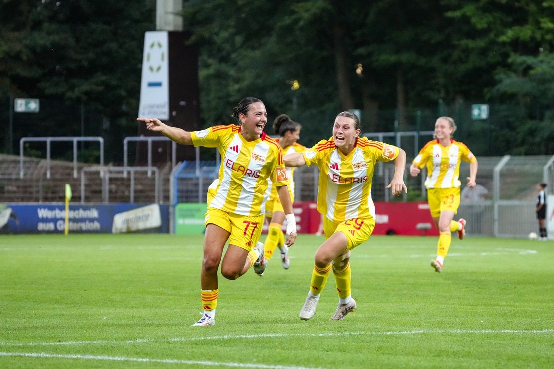 Zwei Fußballspielerinnen in gelben Trikots jubeln auf dem Spielfeld nach einem Tor. Im Hintergrund sind weitere Spielerinnen sichtbar.