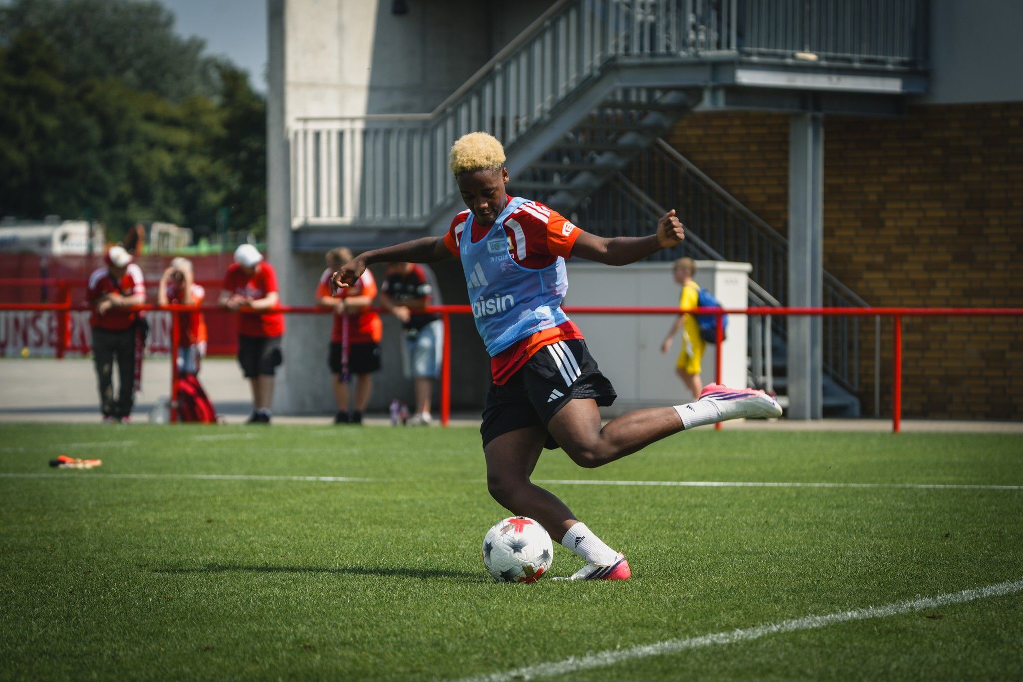 Eine Person mit blonden Haaren in Sportkleidung schießt einen Fußball auf einem Trainingsplatz. Zuschauer im Hintergrund.