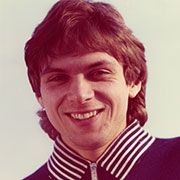 Ein lächelnder Mann mit mittellangen braunen Haaren, der ein blaues Oberteil mit weißen Streifen trägt.