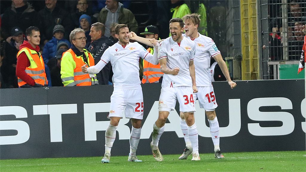 Drei Fußballspieler feiern euphorisch nach einem Tor, dabei sind Zuschauer im Hintergrund zu sehen.