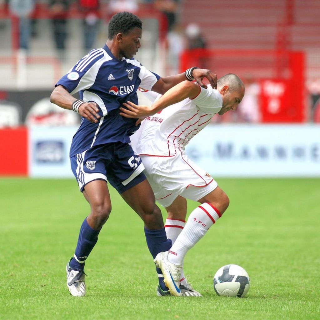 Zwei Fußballspieler in Aktion, einer dribbelt und der andere versucht, den Ball zu erobern. Rasenfeld im Hintergrund.