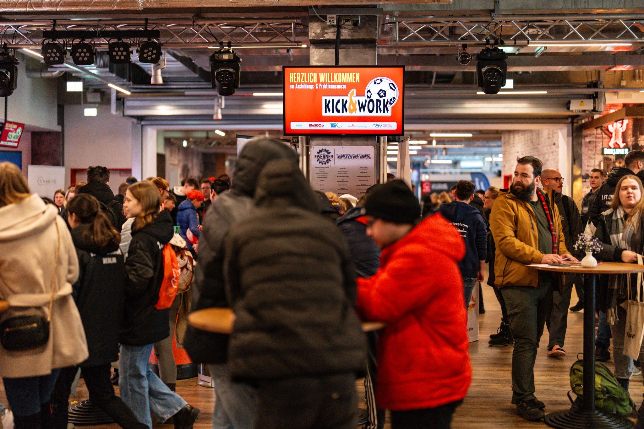 Menschenmenge in einem modernen Veranstaltungsraum mit Ständen, Tischen und einer Begrüßungsanzeige im Hintergrund.