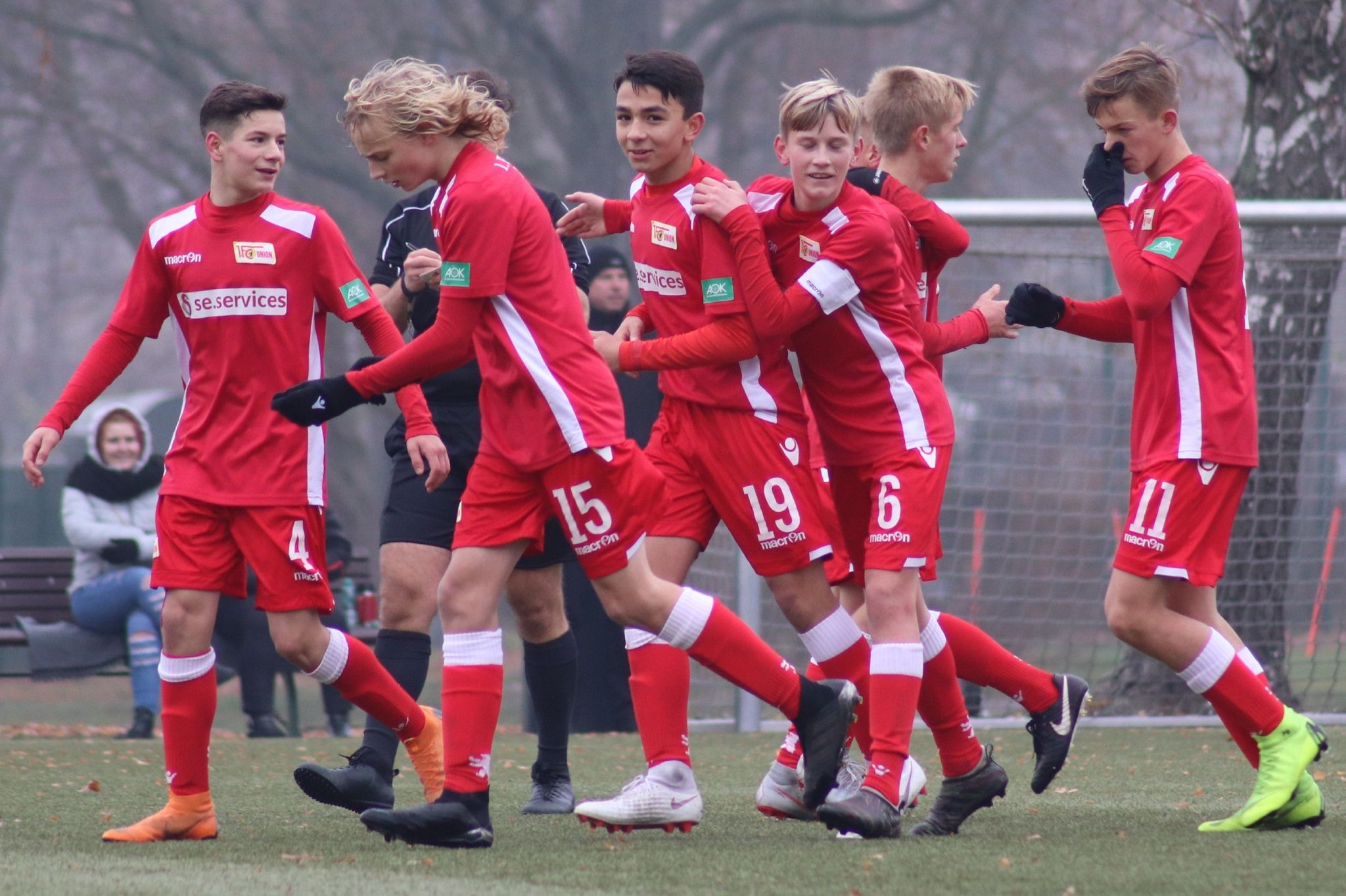 Sechs Fußballspieler in roten Trikots jubeln zusammen auf einem Spielfeld, umgeben von einem unscharfen Hintergrund.