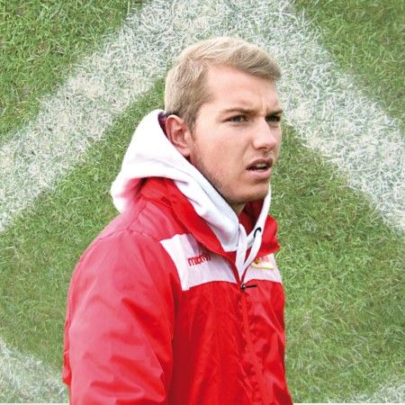 Ein junger Mann in roter Trainingsjacke steht auf einem Fußballfeld mit grünem Rasen und einem weißen Markierungsmuster im Hintergrund.