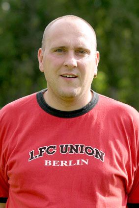 Mann mit Glatze trägt ein rotes Shirt mit dem Schriftzug „1. FC Union Berlin“ vor einem unscharfen natürlichen Hintergrund.