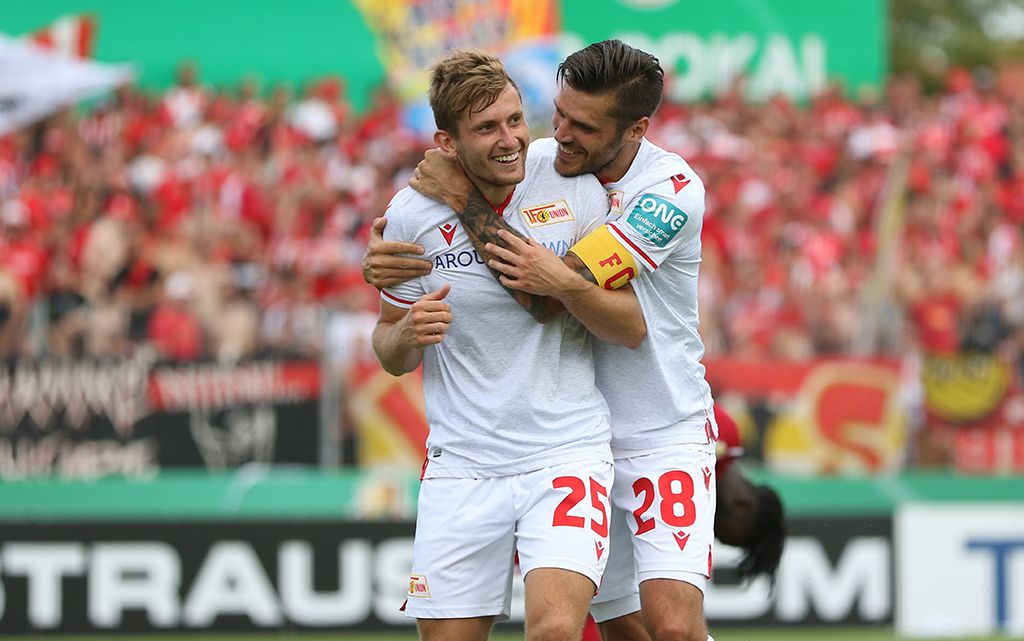 Zwei Fußballspieler umarmen sich freudig auf dem Spielfeld, Hintergrund mit jubelnden Fans in roten Trikots.