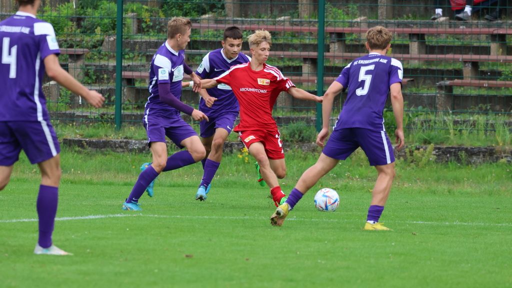 Ein Fußballspiel auf einem Rasenplatz, bei dem ein Spieler in roter Kleidung einen Ball zwischen Gegnern in lila Trikots dribbelt.