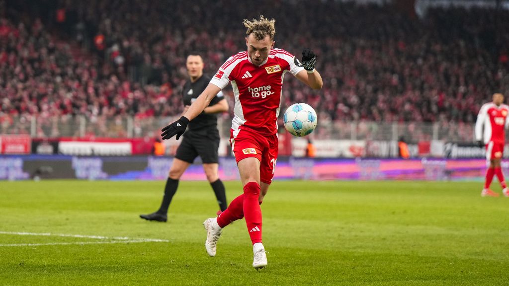 A football player in a red jersey is dribbling the ball during a game in front of a cheering crowd.