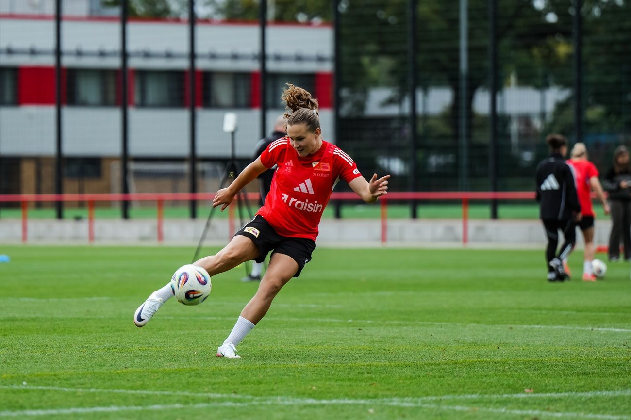 Eine Spielerin in rotem Trikot tritt einen Fußball auf einem Trainingsplatz mit Rasen und Spielern im Hintergrund.