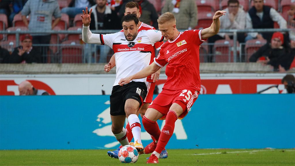 Zwei Fußballspieler kämpfen um den Ball während eines Spiels auf dem Rasen. Im Hintergrund sind Zuschauer im Stadion zu sehen.
