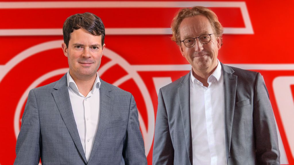 Zwei Männer in Anzügen stehen vor einem roten Hintergrund mit dem Logo eines Unternehmens.