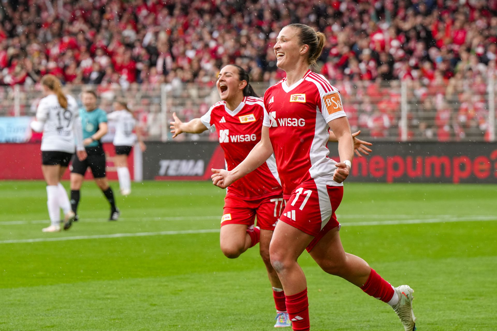Zwei Spielerinnen von Union Berlin in roten Trikots jubeln auf dem Spielfeld. Im Hintergrund sind jubelnde Fans und Spieler zu sehen.